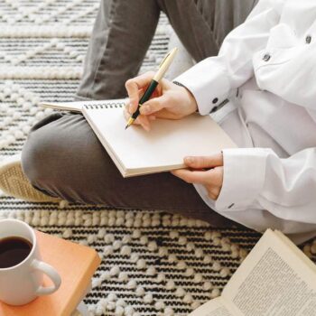 Person sitting cross-legged on a woven rug, journaling about marketing without social media in a notebook, with coffee and books nearby