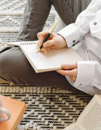 Person sitting cross-legged on a woven rug, journaling about marketing without social media in a notebook, with coffee and books nearby